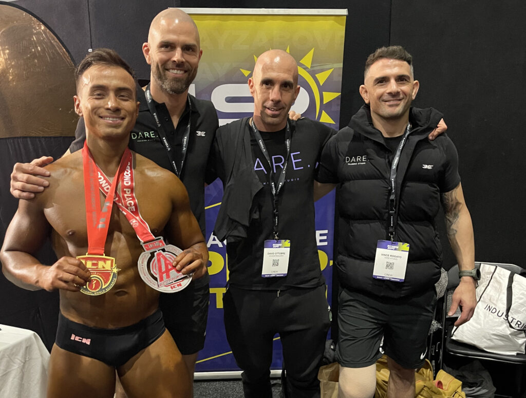 Bodybuilder Seb stands holding a large medal after coming 2nd place at the ICN bodybuilding championships - he stand next to his 3 male personal trainers from Dare Training Studio.